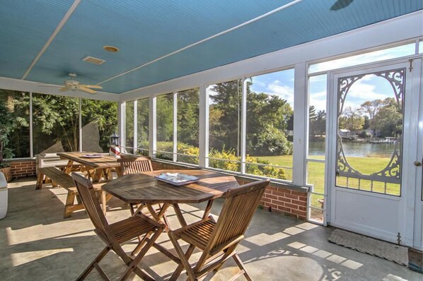 A covered patio with wooden tables and chairs, large windows, and a view of a lake.