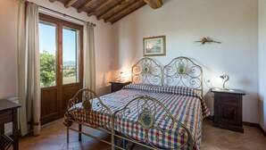 Un dormitori amb un llit de metall, roba de llit amb quadres, un sostre de fusta, una finestra amb cortines, una taula de nit amb una làmpada i un quadre al mur.