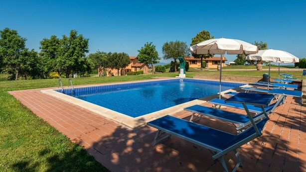 Una piscina rectangular amb vora de rajola, envoltada per un solàrium de maó. Hi ha gandules i ombrelles per prendre el sol.