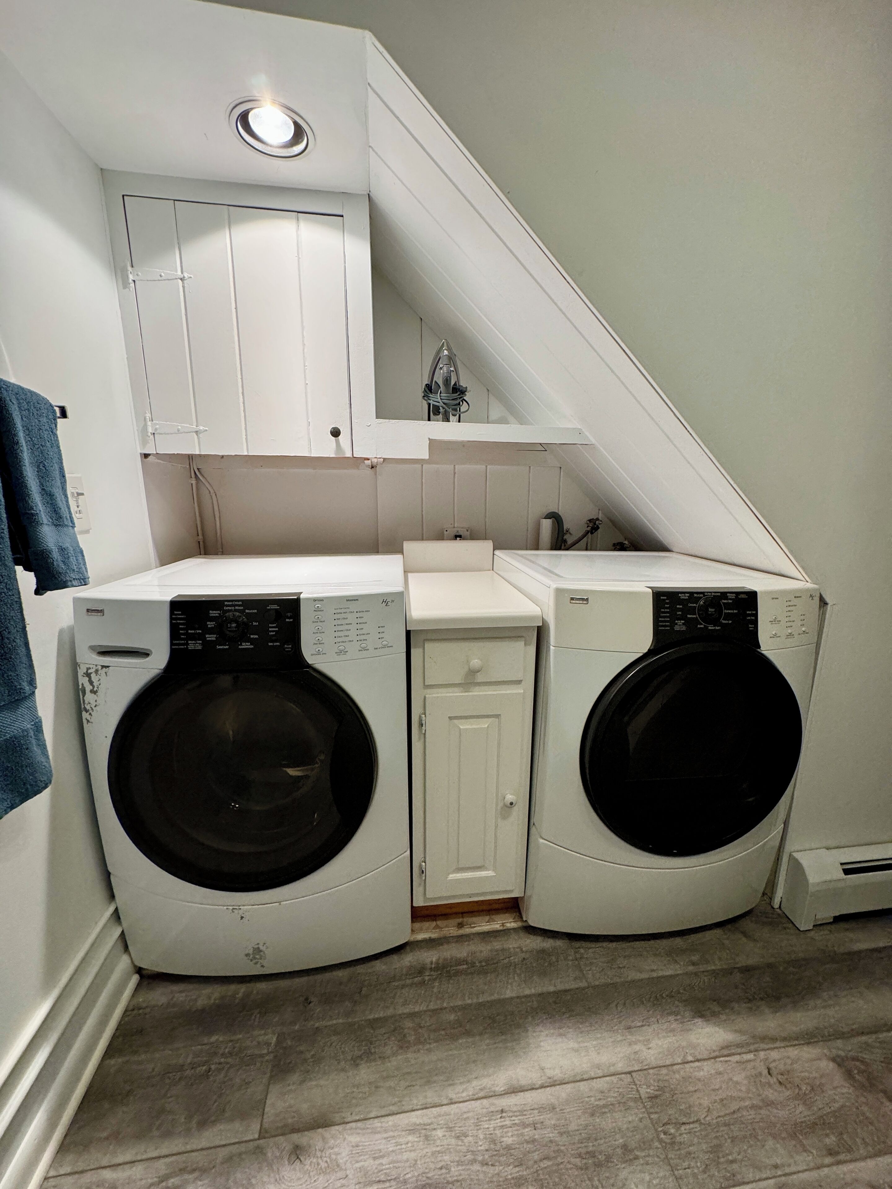 A laundry room with two front-loading washing machines, a small cabinet, and a towel hanging on the wall.
