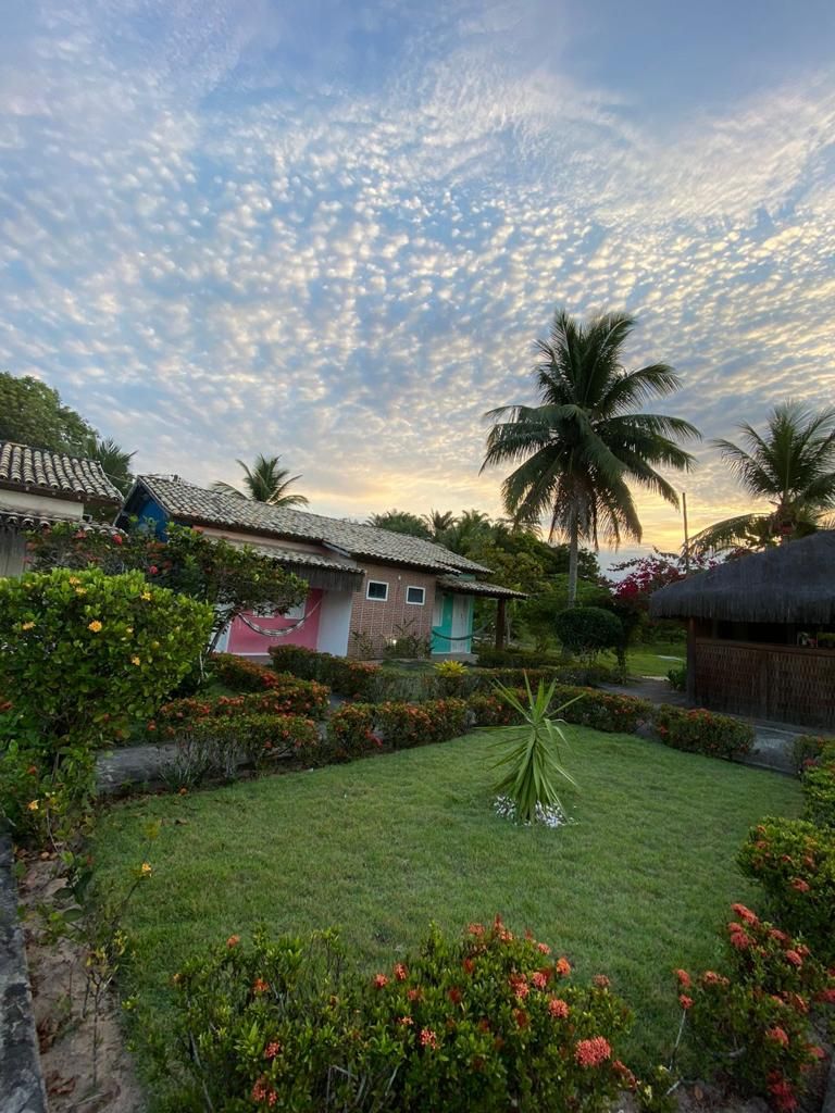 A tropical garden with palm trees, a thatched-roof hut, and a small house with a tiled roof.