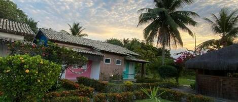 A tropical garden with palm trees, a thatched-roof hut, and a small house with a tiled roof.