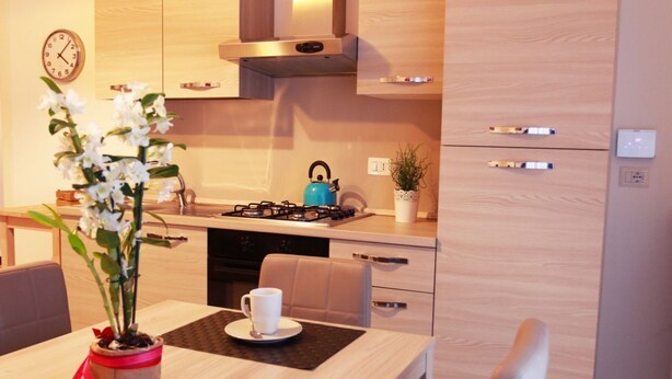 A modern kitchen with wooden cabinets, a gas stove, and a kettle. There is a clock on the wall and a potted plant on the counter. - Bnbook- Via Torino Train Station (Rho)