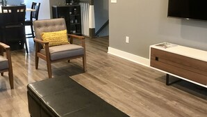 Un salon moderne avec un canapé gris, un pouf noir et une table basse en bois. Un téléviseur à écran plat est fixé au mur, et un escalier mène à un autre niveau.