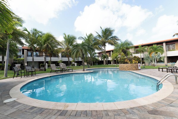 A swimming pool surrounded by palm trees and lounge chairs, with a building in the background. - Eagle Vacation Home (Oranjestad)