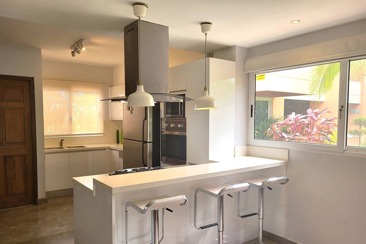 A modern kitchen with a central island, bar stools, stainless steel appliances, pendant lighting, and a window with a view of greenery.