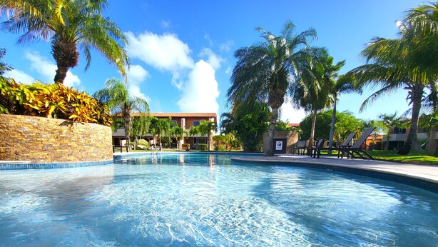 A clear swimming pool surrounded by palm trees and a stone wall, with a building in the background.