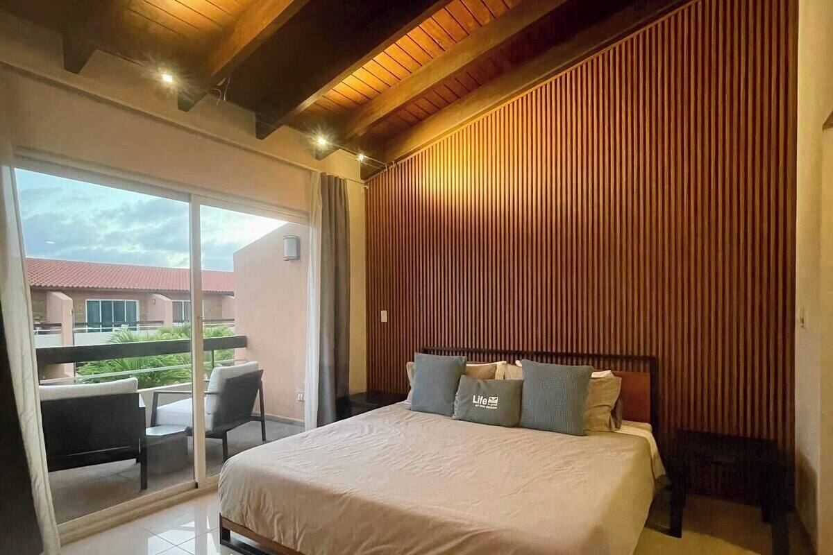 A modern bedroom with a large bed, a wooden slatted headboard, and a balcony with a view of buildings.