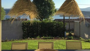 Un espace de restauration en plein air avec une table et des chaises, deux parasols en chaume suspendus et une vue sur un bâtiment et des arbres en arrière-plan.