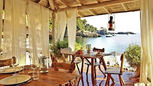 Un espacio de comedor al aire libre con mesas y sillas de madera, cortinas blancas y vistas al mar.