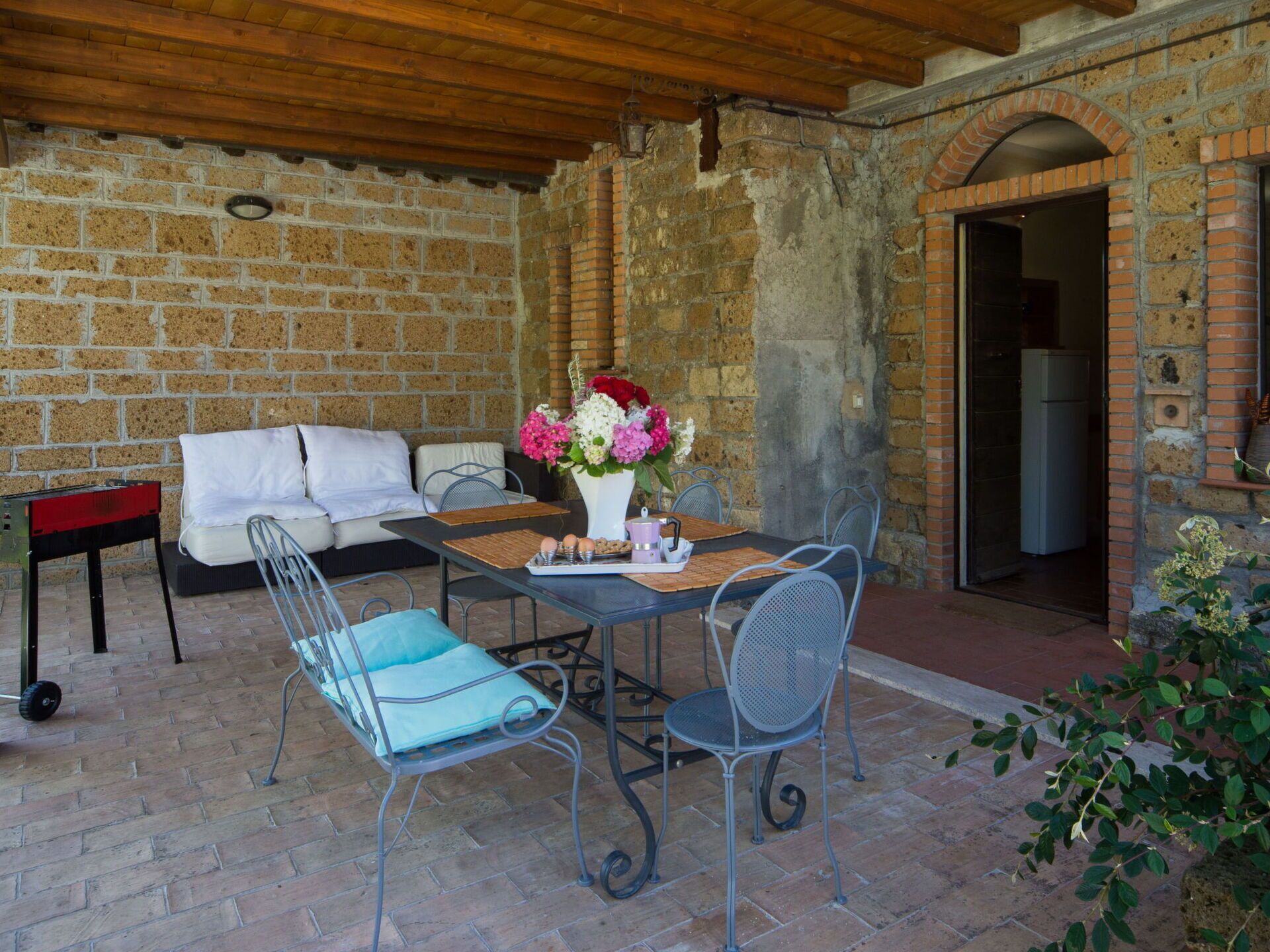 An outdoor patio with a brick wall, a wooden ceiling, a table with chairs, a sofa, and a small table with a vase of flowers.
