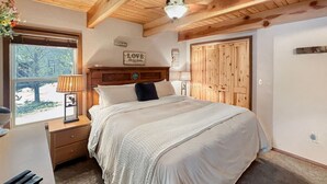 A bedroom with a large bed, wooden ceiling beams, a nightstand with a lamp, and a window with a view of trees.