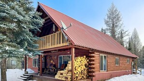 A log cabin with a red roof, a wooden porch, and a stack of firewood outside.