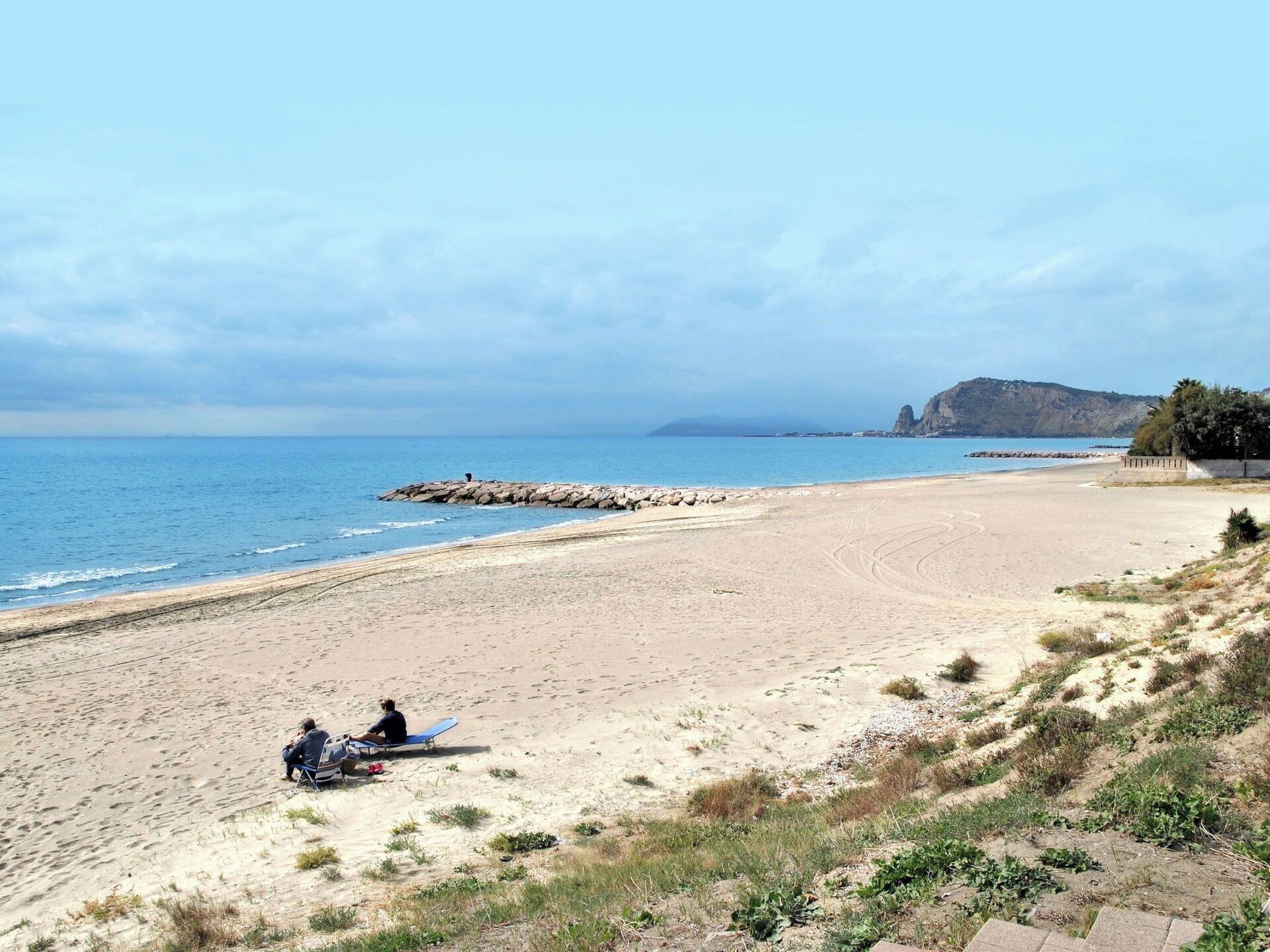 Una playa con un rompeolas rocoso, cielos despejados y una montaña a lo lejos.