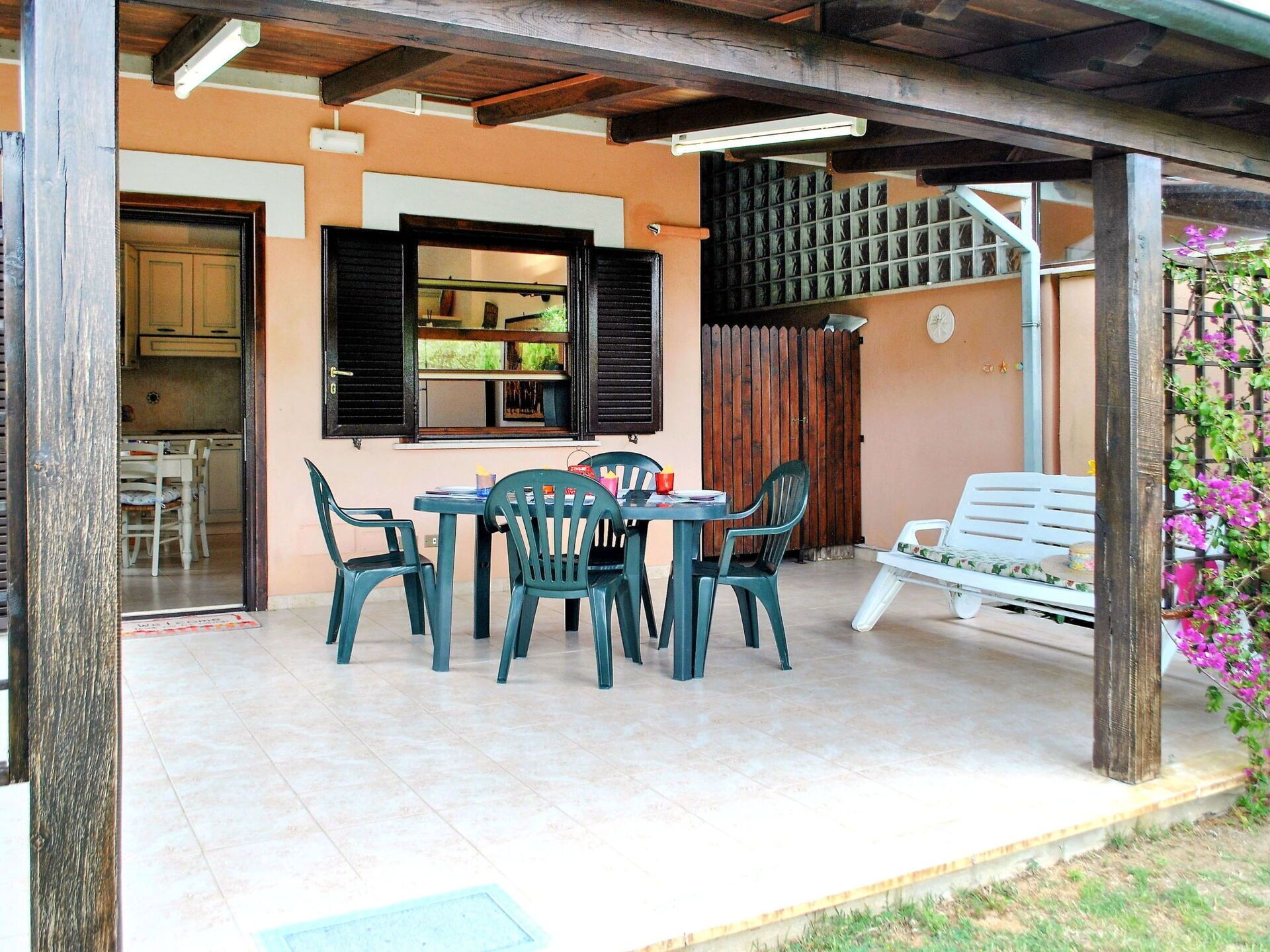 A covered patio with a table and chairs, a bench, and a view of a kitchen and dining area through open shutters.