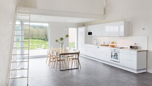 Una cocina moderna con armarios blancos, un comedor con mesa y sillas de madera, y vistas a un paisaje verde a través de amplios ventanales.