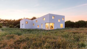 Una casa moderna de dos pisos con techo plano y varias ventanas, rodeada por un campo cubierto de césped y árboles.