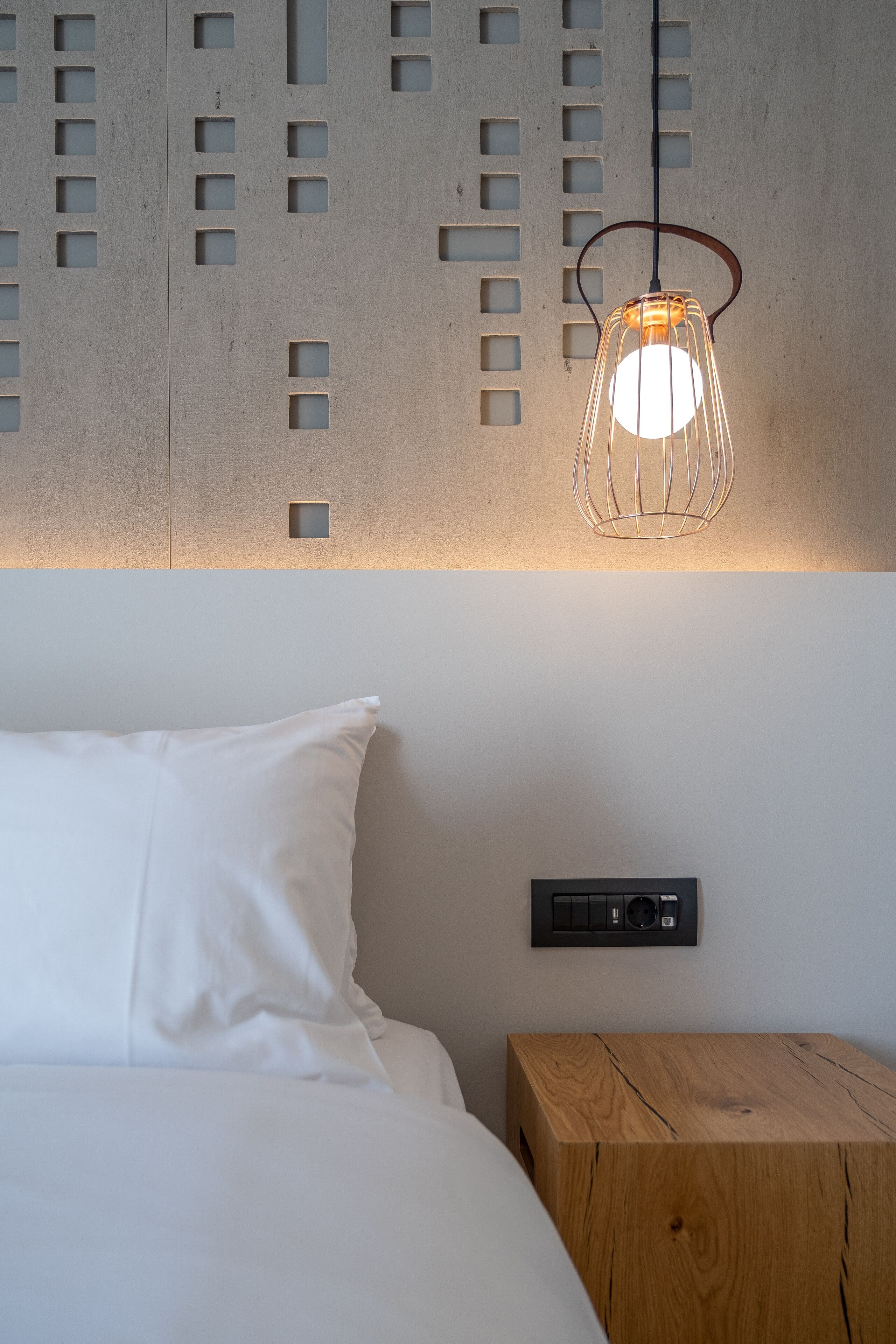 A bedroom with a bed, a bedside table, a wall-mounted light fixture, and a wall panel with cut-out windows.