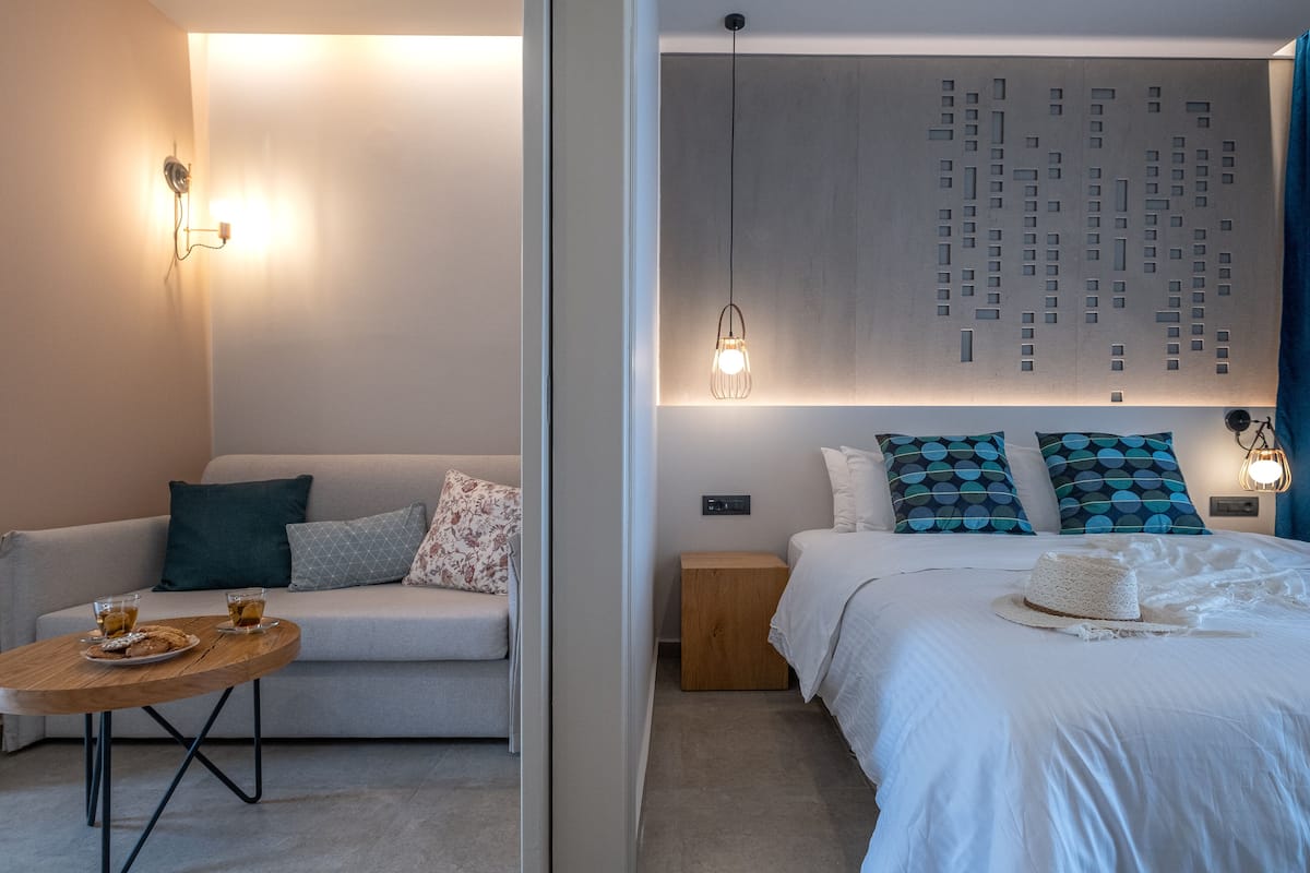 Una habitación de hotel moderna con un sofá, una pequeña mesa de madera con bebidas, una cama con almohadas estampadas y una lámpara fijada a la pared.