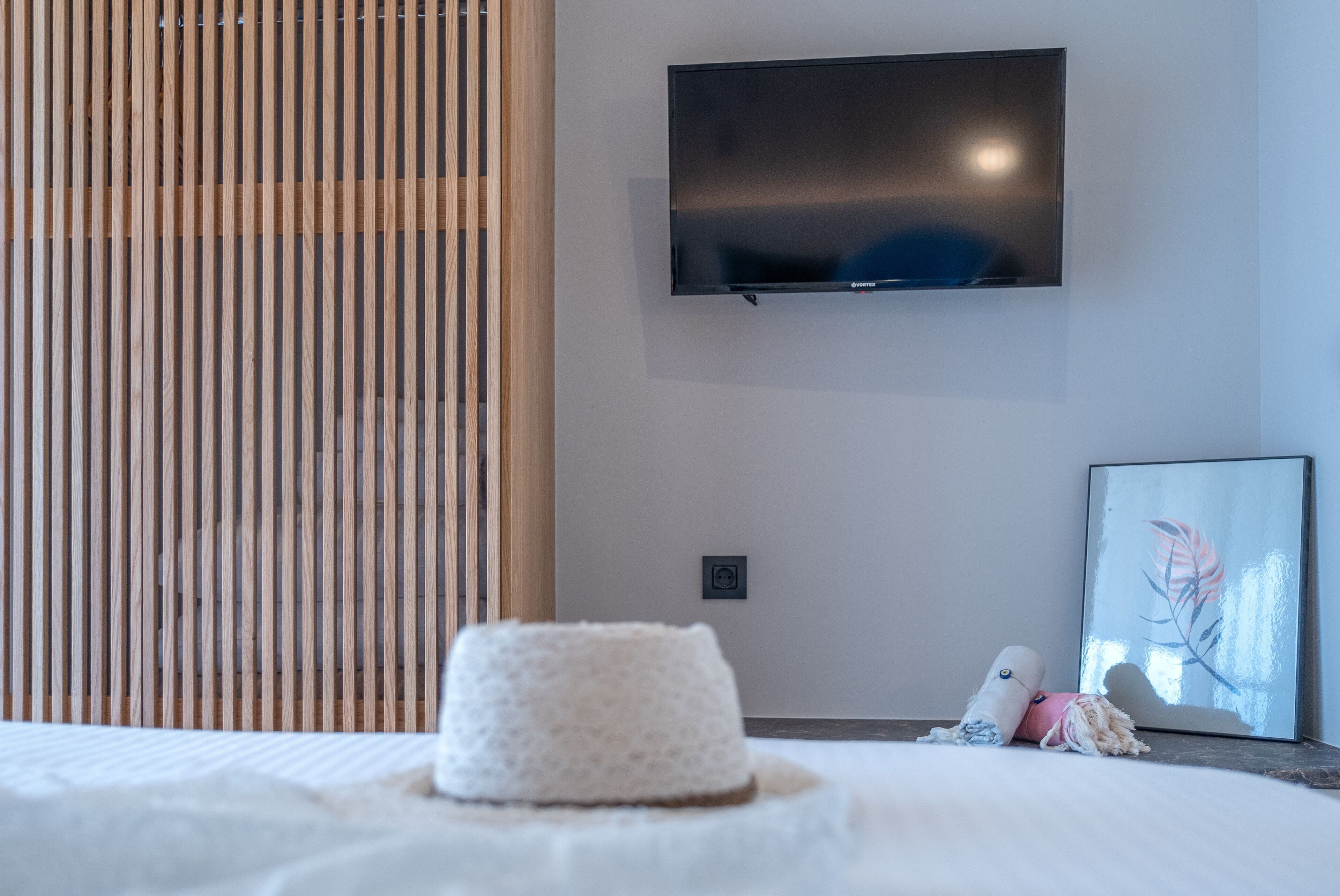 A modern hotel room with a flat-screen TV mounted on the wall, a wooden slat window, a bed with a white headboard, and a framed artwork on the wall.