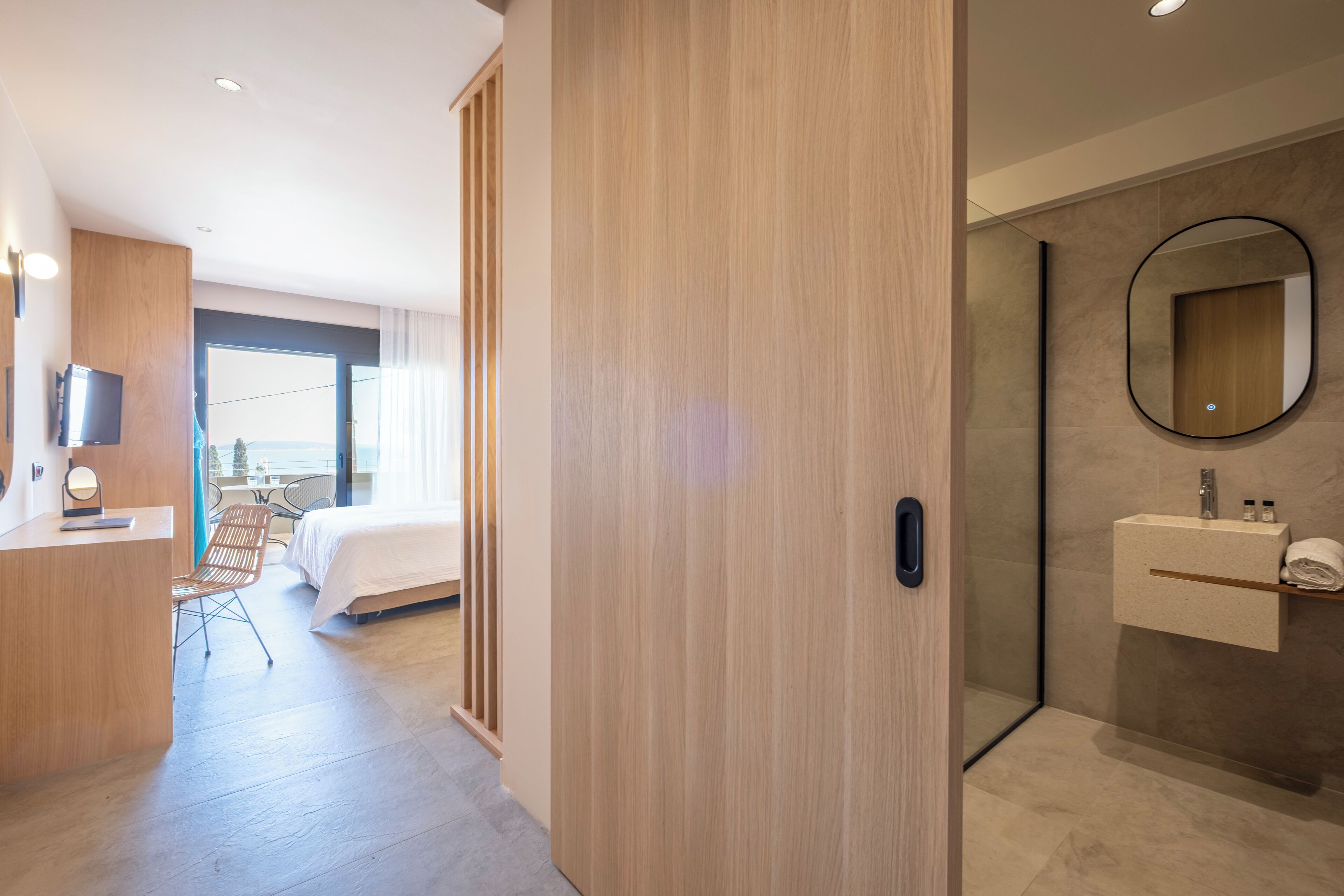 Een moderne hotelkamer met een houten garderobe, een bed, een bureau met stoel, een badkamer met spiegel en wastafel, en een groot raam.