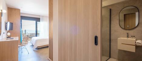 A modern hotel room with a wooden wardrobe, a bed, a desk with a chair, a bathroom with a mirror and sink, and a large window.