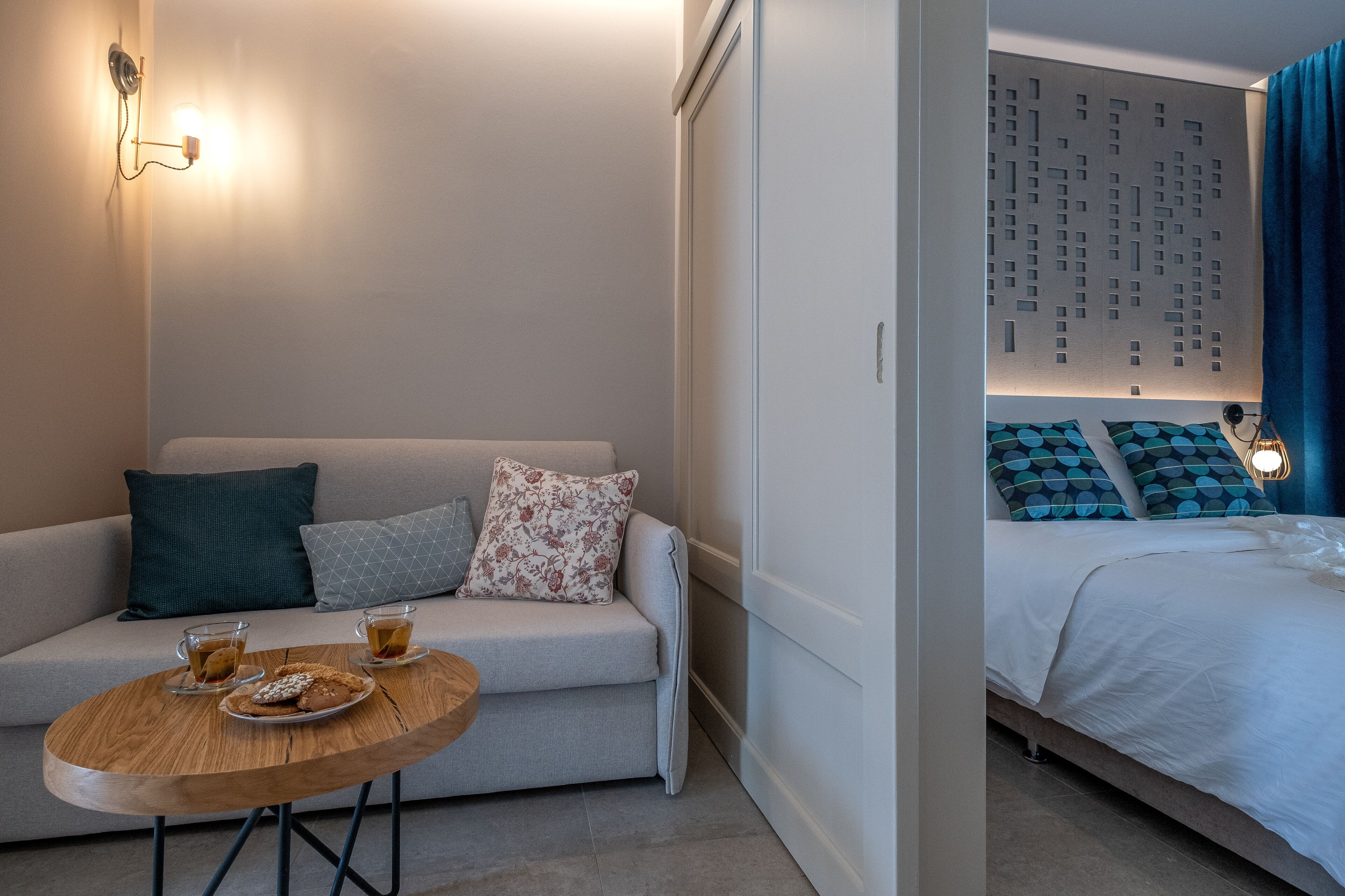 A modern hotel room with a sofa, a small round table with drinks and cookies, a bed with patterned pillows, and a wall with a geometric design.