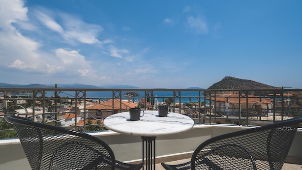 A balcony with a round table and two chairs, overlooking a coastal town with buildings and a mountain in the distance. - Elixia (Nafplio)