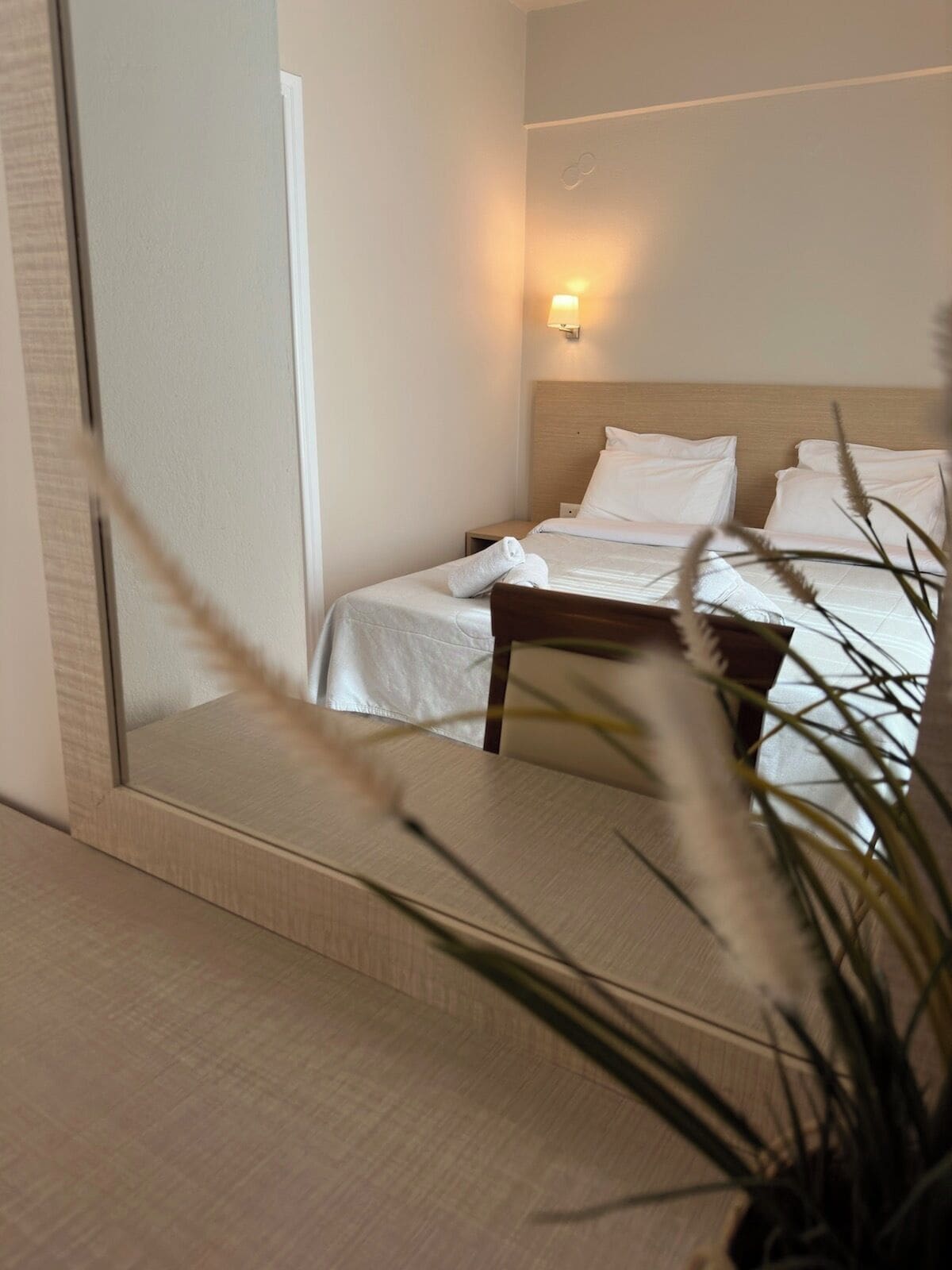 Una habitación de hotel con una cama, una mesita de noche, una lámpara fijada a la pared y un espejo que refleja parte del cuarto.