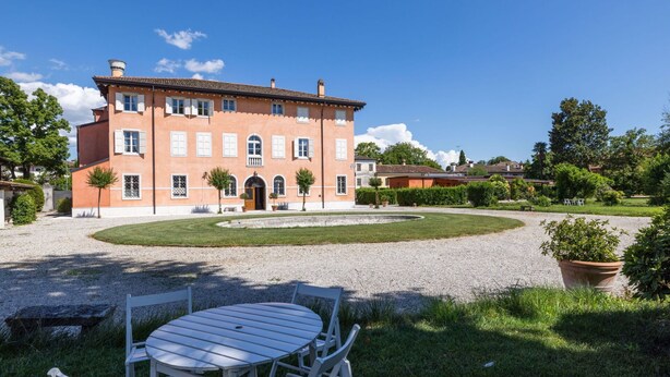Un grand bâtiment à deux étages, doté d’une façade symétrique, d’une entrée centrale surmontée d’un portique, et d’un jardin bien entretenu avec un chemin de gravier et une table ronde entourée de chaises, le tout mis en valeur au premier plan.