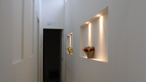 A narrow, well-lit hallway with a door at the end, a wall-mounted shelf with a potted plant, and recessed lighting. - MODERN HOUSE 4 SUITES WITH POOL IN GERIBA (Campo de Pouso)