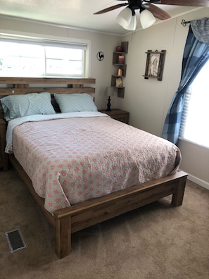 A bedroom with a wooden bed, a ceiling fan, a window with curtains, and a nightstand with a lamp.