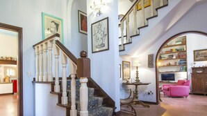 Un magnifique escalier, orné de balustrades travaillées et recouvert d’un tapis à motifs, mène à un palier. Un lustre est suspendu au-dessus, et on aperçoit, au fond, une bibliothèque remplie de livres et d’objets décoratifs.