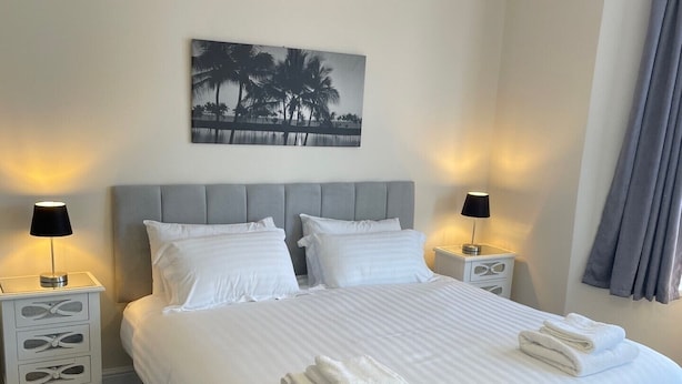 Un lit bien fait, avec du linge de lit et des serviettes blanches, deux tables de chevet avec des lampes, une tête de lit et un tableau représentant des palmiers accroché au mur.