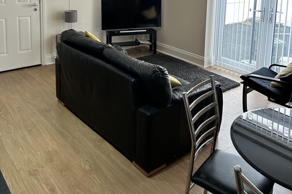 A living room with a black sofa, a TV on a stand, a chandelier, and a dining area with a table and chairs.