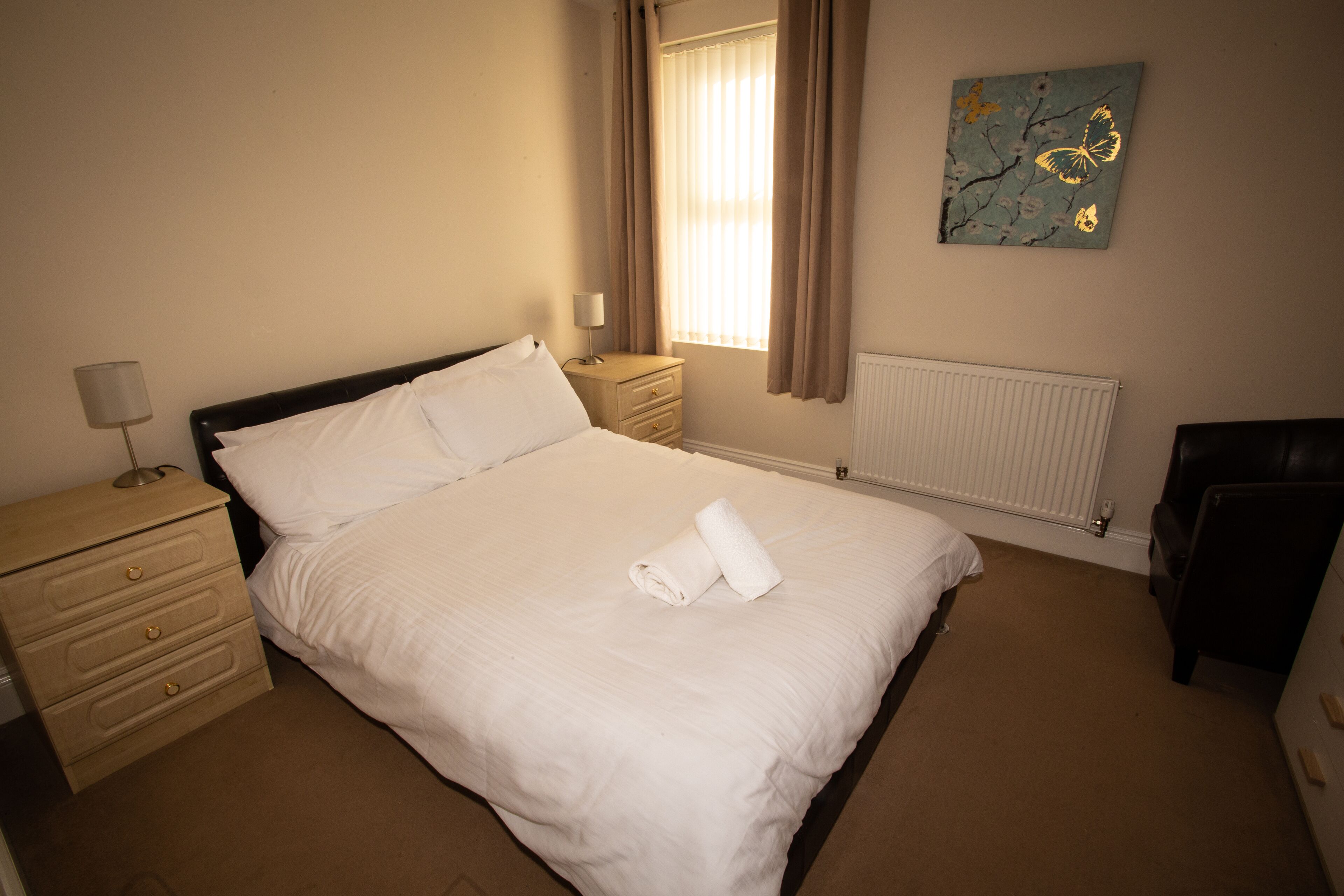 A neatly made bed with white linens, a bedside table with a lamp, a radiator, and a painting on the wall.