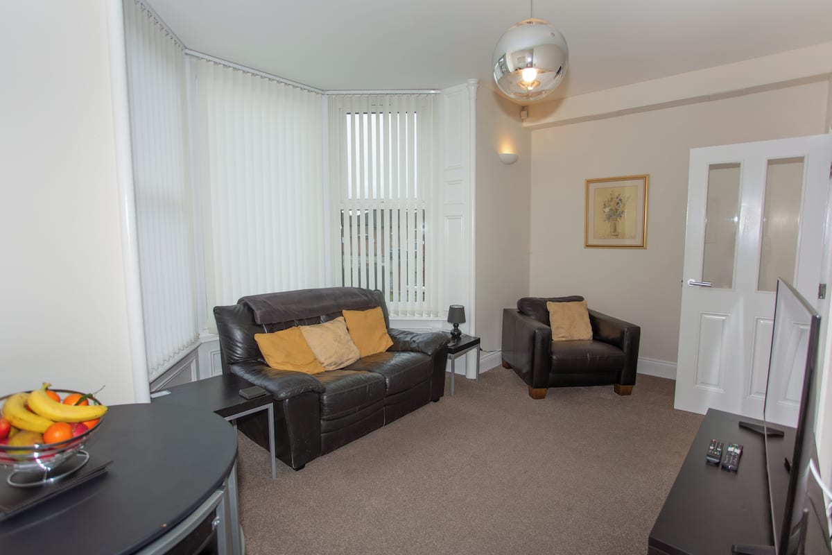 A living room with a black leather sofa, a smaller armchair, a glass coffee table with fruit, and a TV on a stand.