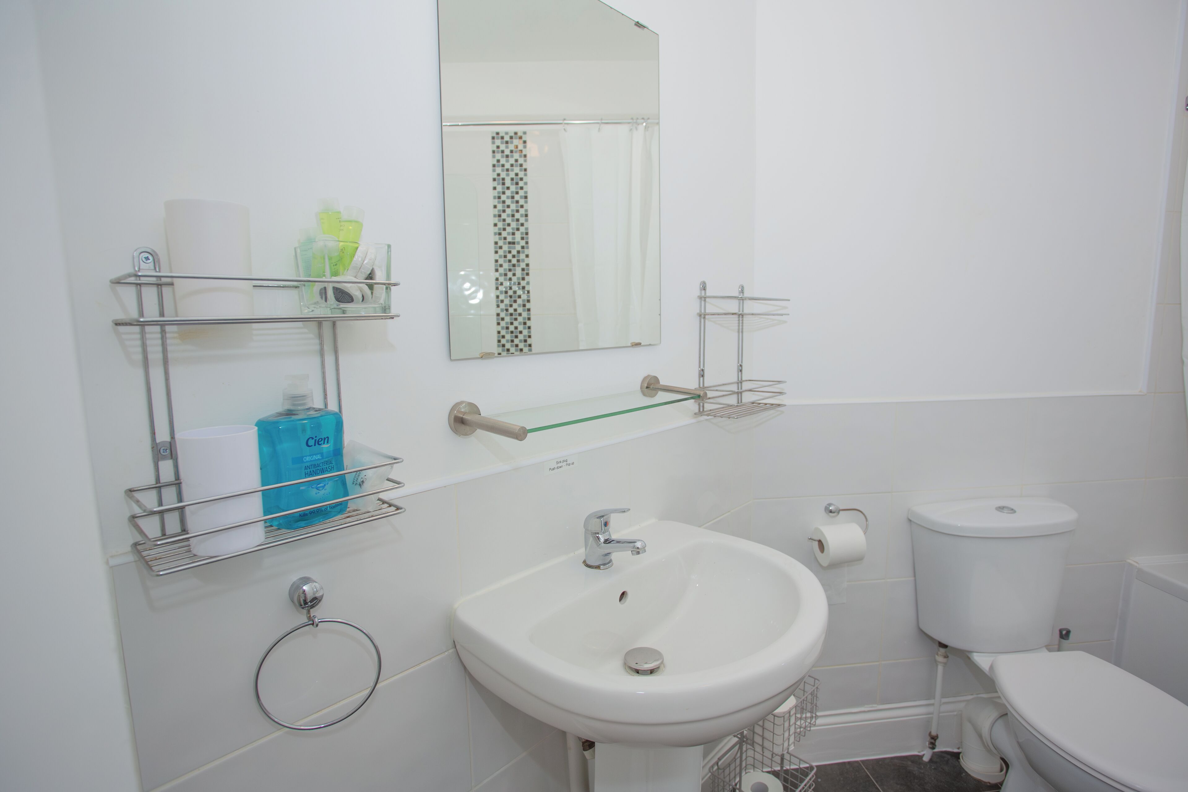 A modern bathroom with a white sink, toilet, and mirror. There are shelves with toiletries and a towel ring.