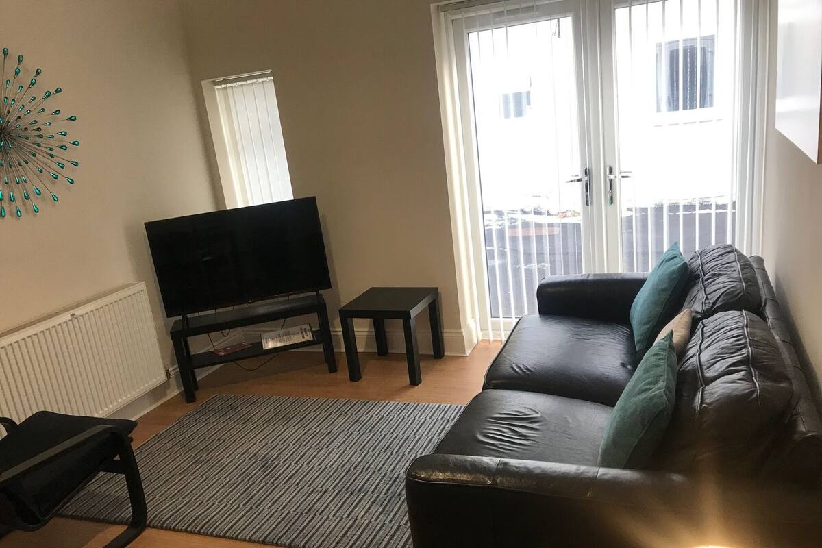 A living room with a black leather sofa, a television on a stand, a small table, and a patterned rug.