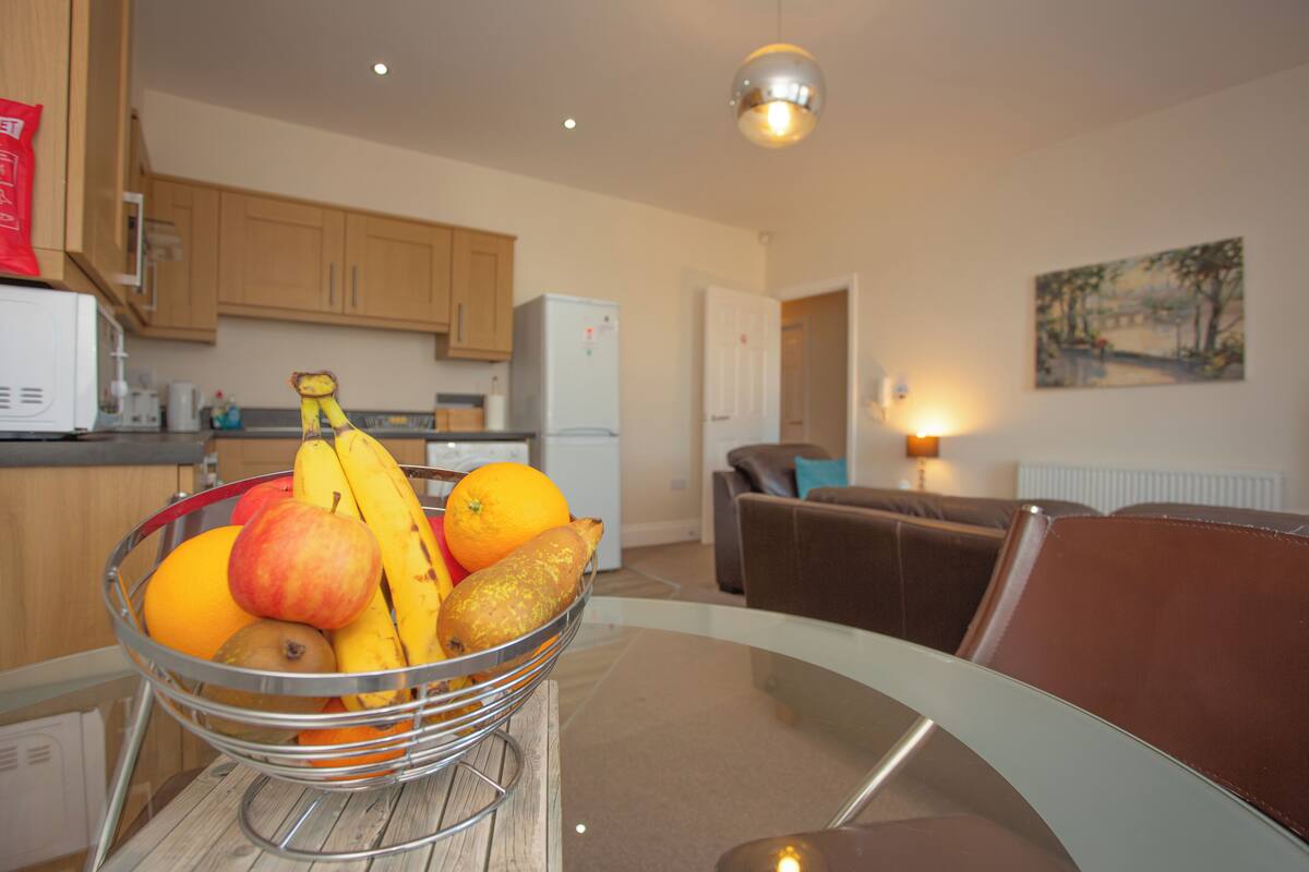 A kitchen with wooden cabinets, a refrigerator, and a fruit bowl on a glass table.