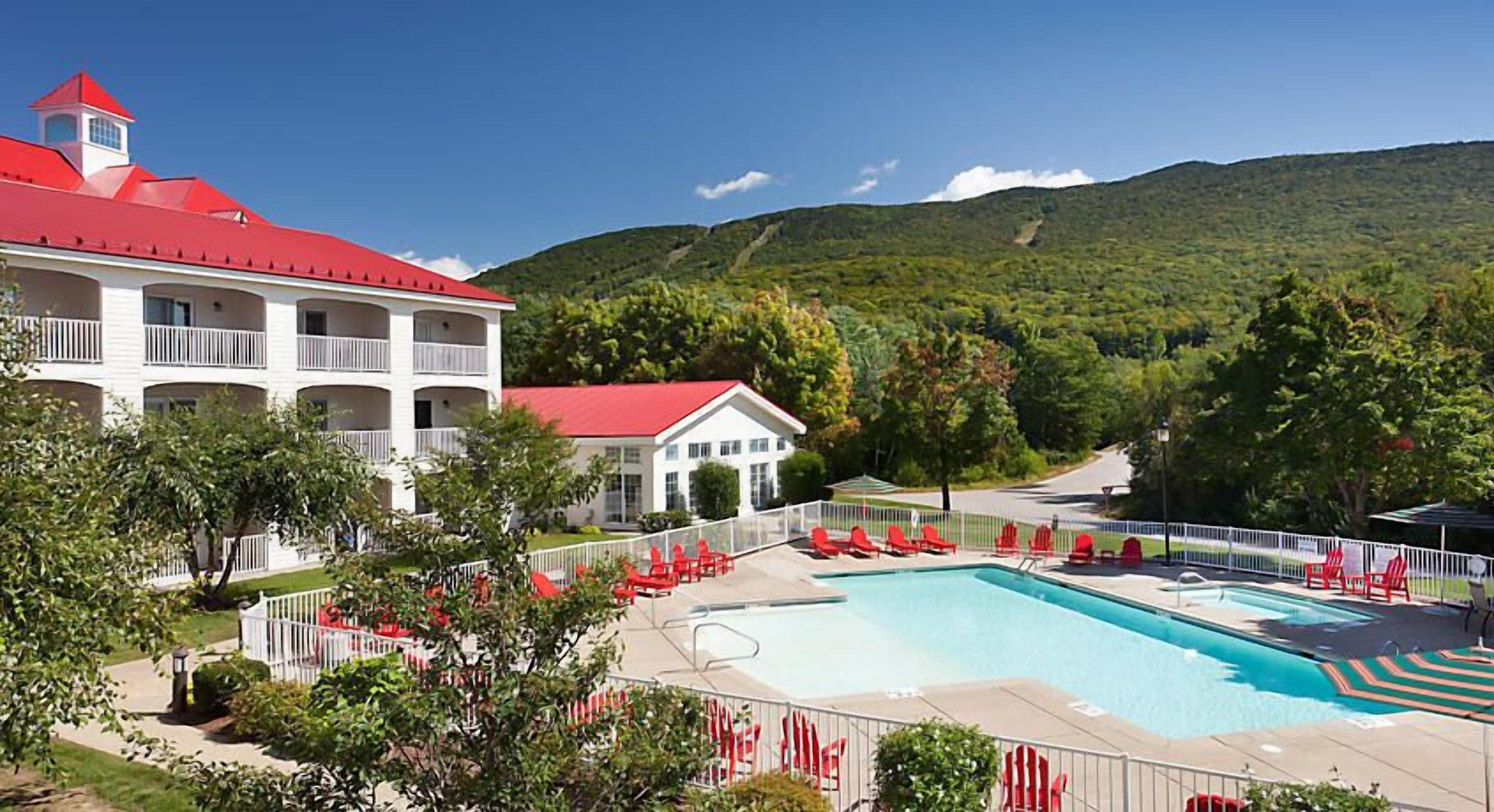 A hotel with a swimming pool, red chairs, and a view of a green mountain.