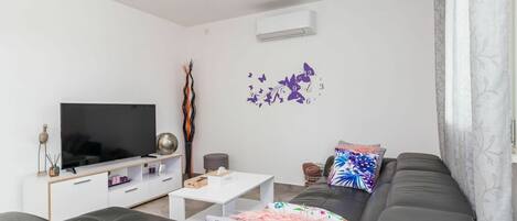 A modern living room with a flat-screen TV, a white coffee table, a grey sofa with patterned cushions, and a wall-mounted air conditioner.