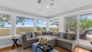 A spacious living room with a large sectional sofa, a coffee table with flowers, and a view of a natural landscape through large windows. - Gorgeous views!  Charming home on the river (Kiawah Island)