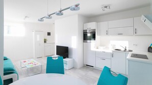 A modern kitchen with white cabinets, a built-in oven, and a sink. There is a dining area with a white table and teal chairs.