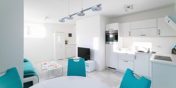 A modern kitchen with white cabinets, a built-in oven, and a sink. There is a dining area with a white table and teal chairs.