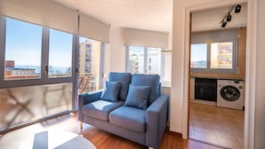 A modern living room with a grey sofa, large windows, a washing machine, and a view of buildings. - HomeHolidaysRentals Elodie - Costa Barcelona (Calella)