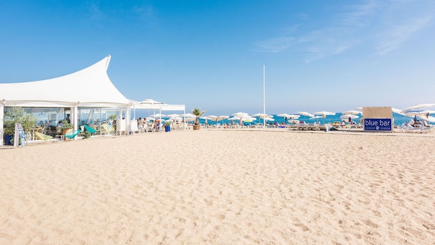 A beach with a white tent structure, umbrellas, and a 'blue bar' sign. - HomeHolidaysRentals Elodie - Costa Barcelona (Calella)