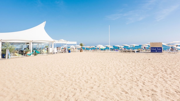A beach with a white tent structure, umbrellas, and a 'blue bar' sign.