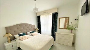 A bedroom with a tufted headboard bed, bedside tables with lamps, a dresser, a mirror, and a television on the wall.