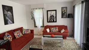 A living room with two red sofas, a coffee table, and a black chair. There are two framed artworks on the walls, a floor lamp, and a TV on the right.